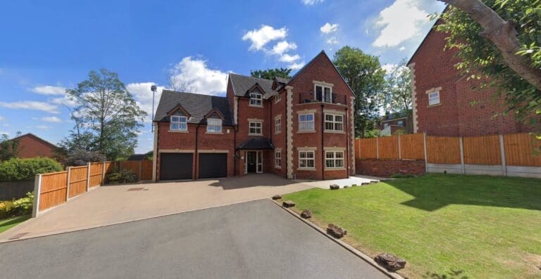 A large red brick house with multiple floors and a double garage, surrounded by a paved driveway and a grassy lawn, under a blue sky with clouds.